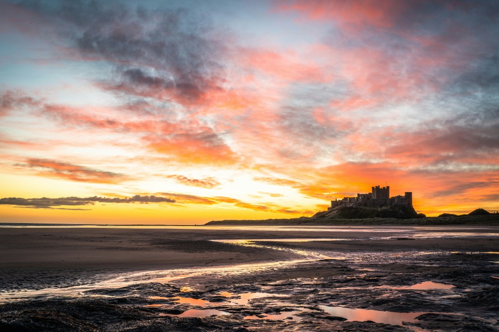View of Bamburgh Castle at sunrise, with colourful clouds and reflections on the wet sand of Bamburgh Beach.